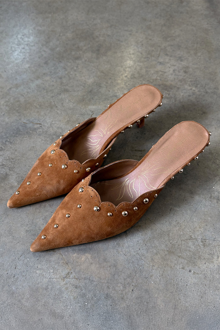 Tan suede Scallop City Mules with gold stud detailing and scalloped edges on a grey stone floor.