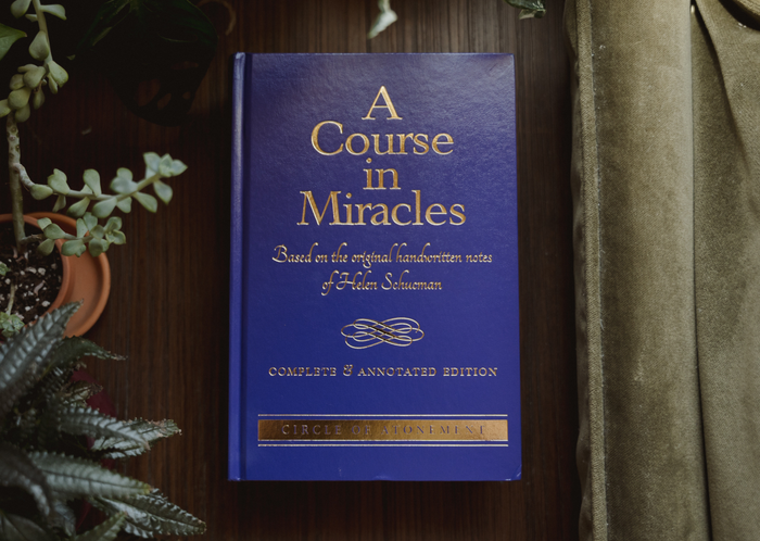 Front cover of A Course in Miracles Complete and Annotated Edition hardcover book, displayed flat on a dark wooden surface surrounded by green indoor plants and curtains.