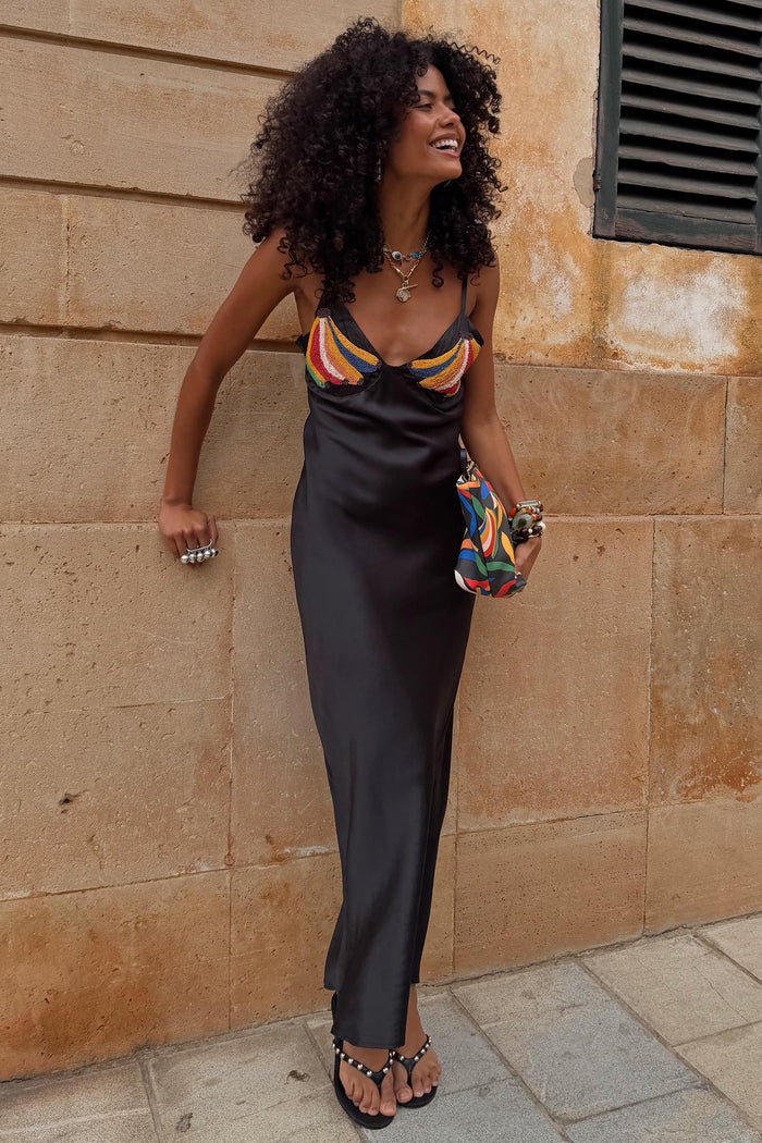 Black Banana Slip Dress with colorful beaded banana motifs, bias-cut silhouette, and V-neckline against a stone wall.