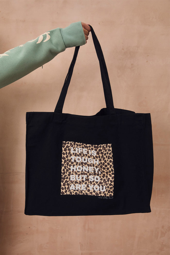Black canvas Life Is Tough Tote Bag featuring a leopard print patch and motivational slogan, held against a beige wall.
