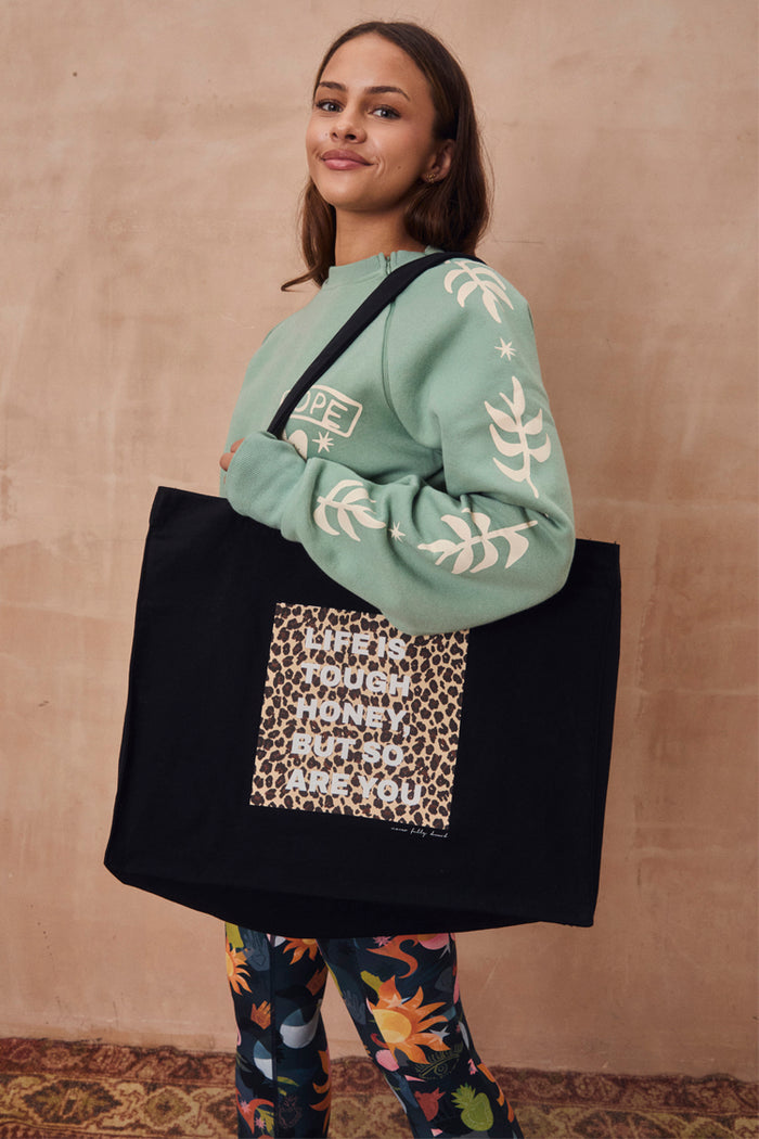 Black canvas Life Is Tough Tote Bag featuring an iconic leopard print slogan graphic, styled over the shoulder.