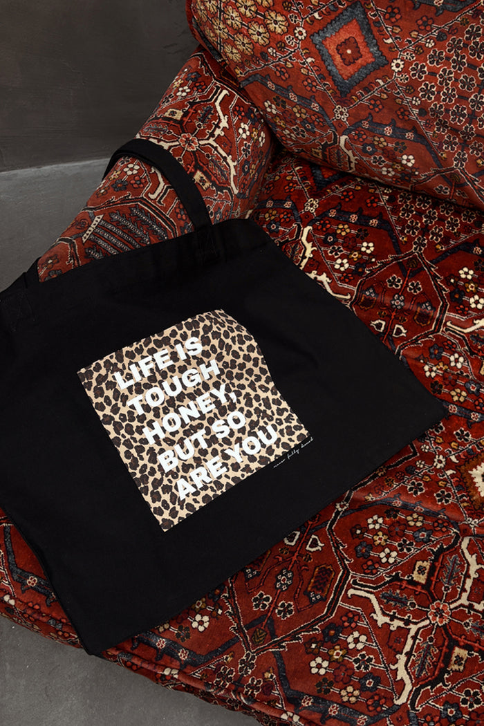 Black canvas tote bag with leopard print 'Life Is Tough Honey' graphic, resting on a patterned armchair.
