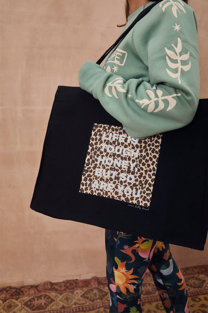 Black canvas tote featuring iconic leopard print and motivational text, carried over the shoulder by a model.