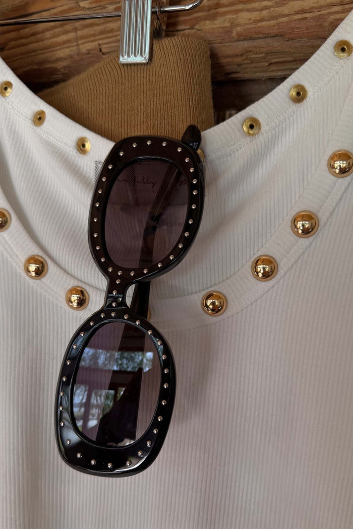 Black Studded Acetate Sunglasses with silver studs and dark lenses, hooked onto a white ribbed top.