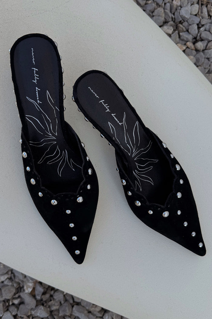Pointed black suede mules with silver studs and scalloped edging, shown on a neutral stone background.