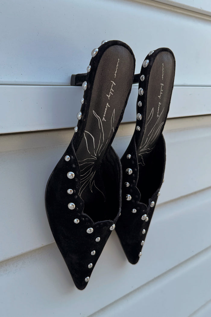 Black suede mules with silver stud detailing and scalloped edges, leaning against a white slatted background.