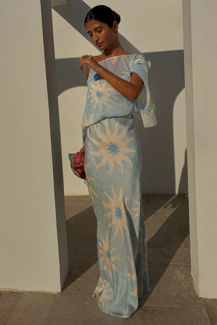 NFD Blue Soleil Anais Dress with a Mediterranean sun print, featuring a draped off-the-shoulder top and flowing skirt.