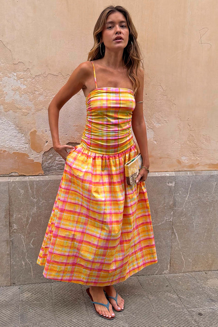 Check Lola Mid-axi Dress in yellow, pink and orange with a ruched bodice and cotton skirt against a rustic wall.