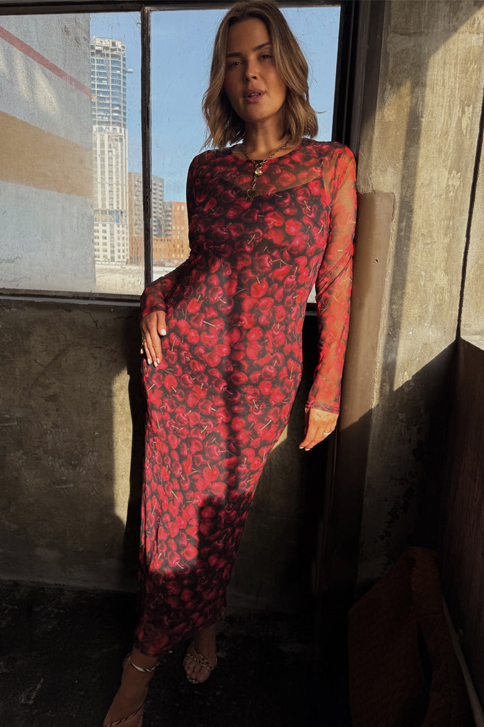 Red Cherry Mesh Soho Dress with a digital cherry print and long sleeves, worn by a model in an industrial setting.