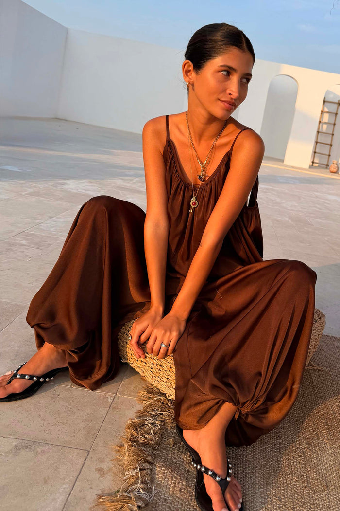 Chocolate Balloon Jumpsuit with a draped silhouette and gathered neckline, shown on a model sitting on a woven stool.