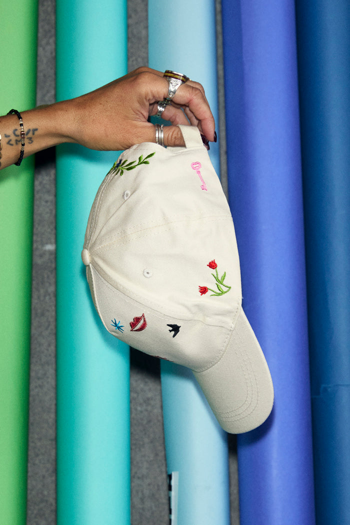 Cream Choose Love Cap with embroidered icons, held by a hand against a backdrop of colourful paper rolls.