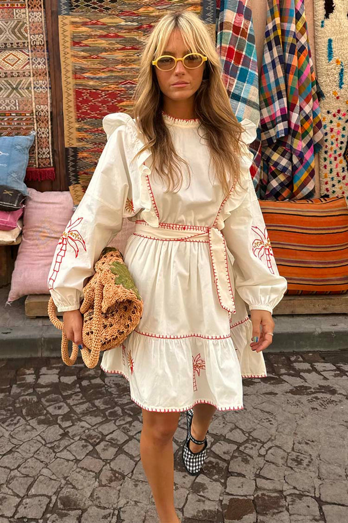 Cream cotton Pollenca Mini Dress with red palm tree embroidery, ruffled shoulders and a tie-waist in a market.