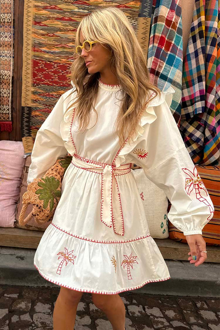 Cream cotton Pollenca Mini Dress with red palm tree embroidery, ruffled shoulders and a belted waist in a vacation setting.