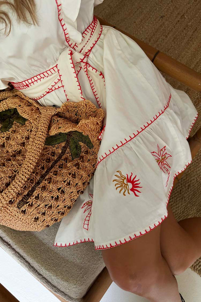 Pollenca Mini Dress in cream cotton with red sun embroidery and a tiered skirt, paired with a woven palm bag.