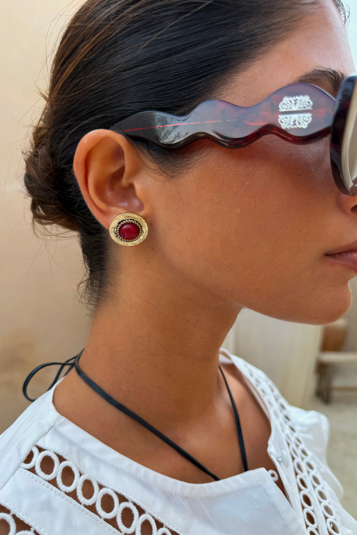 Deloris Red Circle Earrings in textured brushed brass with a red stone, shown on a model wearing tortoiseshell sunglasses.