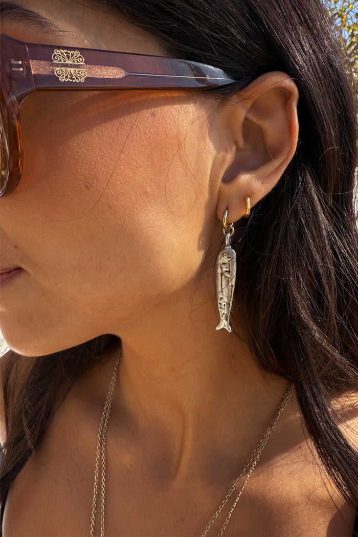 Mixed metal Deloris Sardine Earrings with a silver fish pendant on a gold hoop, shown on a model.