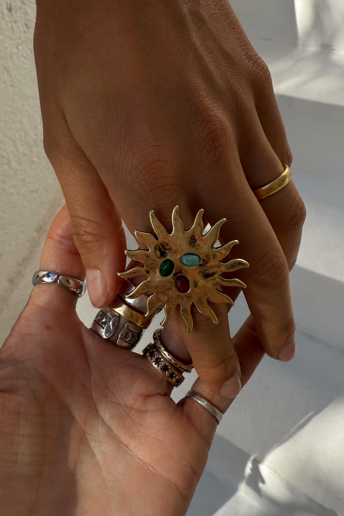 Deloris Sun Ring in brass featuring a sun motif and semi-precious stones, styled with layered jewellery on a hand.