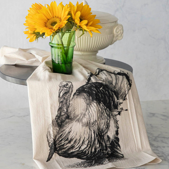 A beige cotton tea towel with a screen-printed black turkey design, displayed on a kitchen counter with a vase of sunflowers in the background.