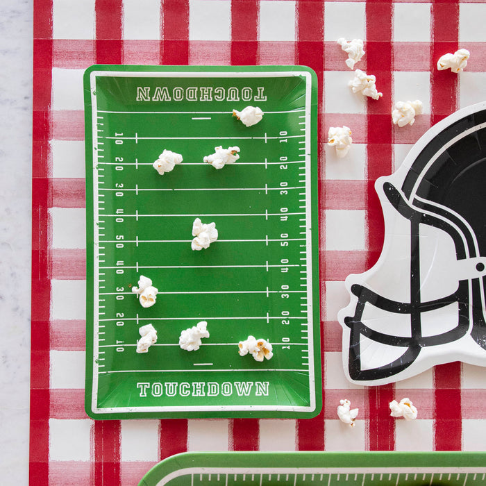 A football field with Football Field Paper Plates and a football helmet on a red and white checkered tablecloth, accompanied by game day goodies.