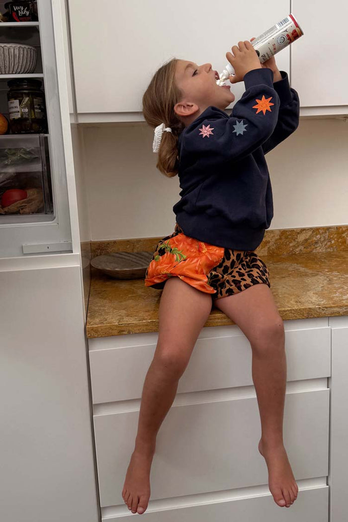 Kids Navy Charms Sweatshirt with colourful star motifs on the sleeves, worn by a young girl in a kitchen setting.
