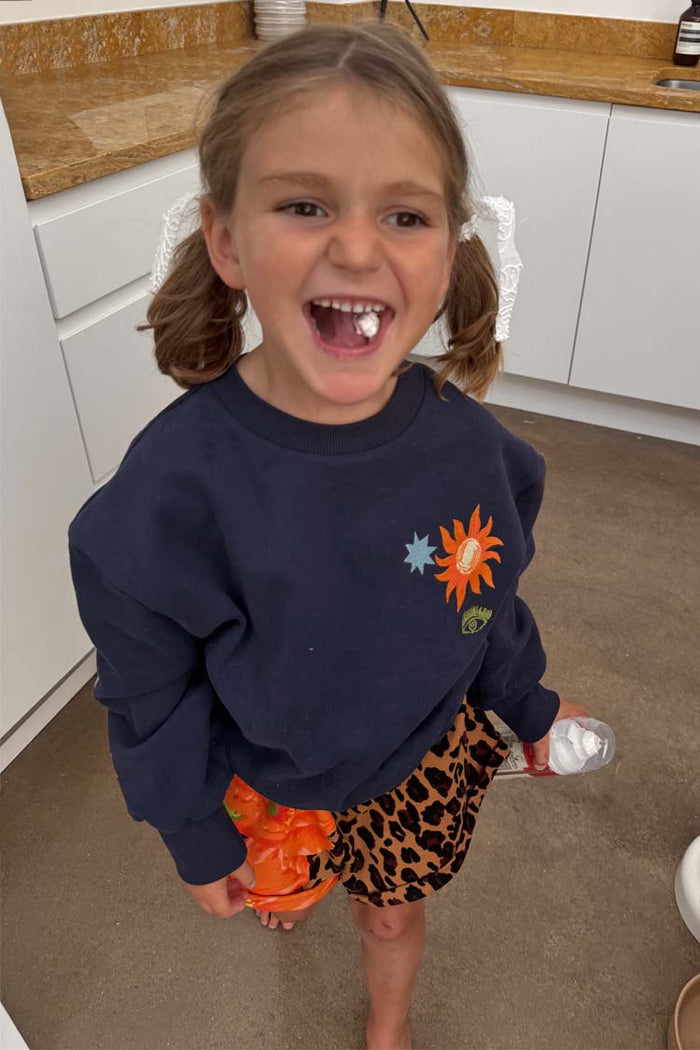 Kids Navy Charms Sweatshirt featuring colourful sun and star motifs, worn by a laughing girl in pigtails.