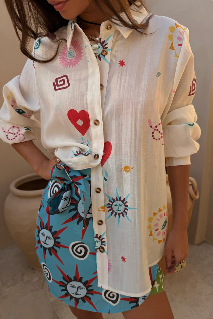 Sheer cream Morocco Charms Marley Shirt with heart and sun motifs, styled with a vibrant blue printed wrap skirt.
