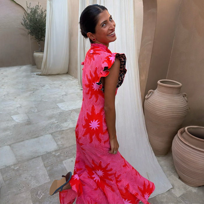 Pink Soleil Remi Dress with red sun motifs and leopard-lined pleated frills on a smiling woman in a courtyard.