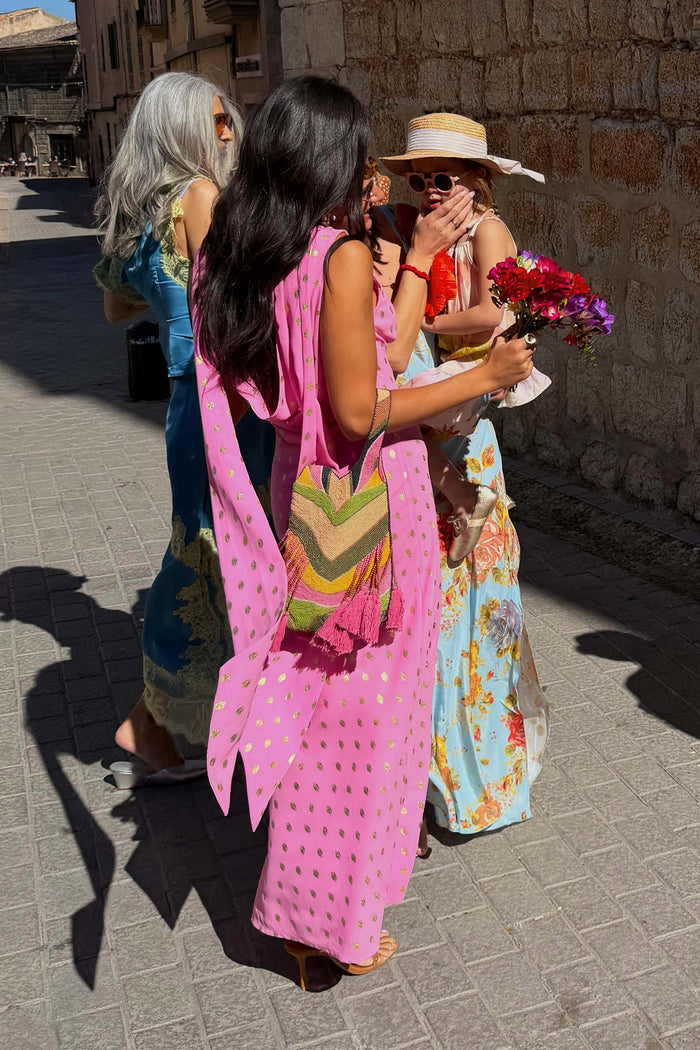 Pink Sophie Dress featuring gold flecks and a multi-wear scarf, styled with a beaded bag on a sunny cobbled street.