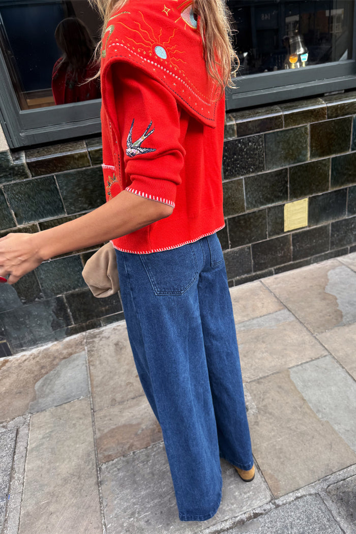 Red Mystical Cardigan with swallow jacquard motifs and blanket stitch detailing, styled with blue wide-leg jeans.