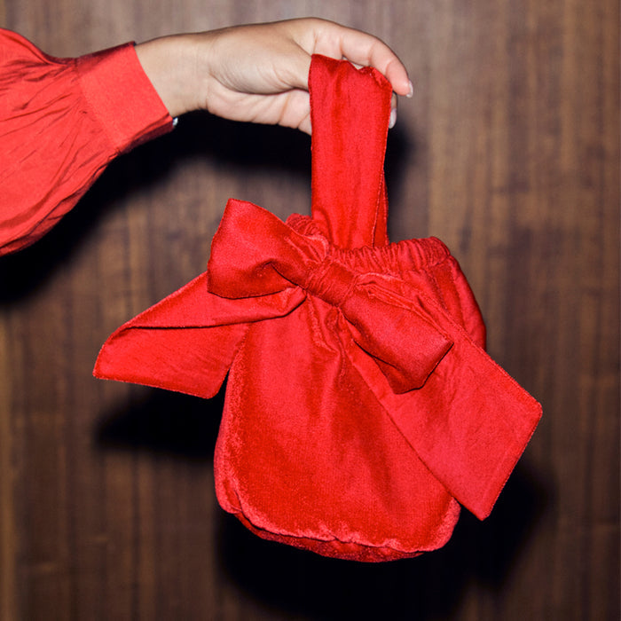 Red Velvet Bow Bag with a large statement bow and top handle, held against a wooden backdrop.