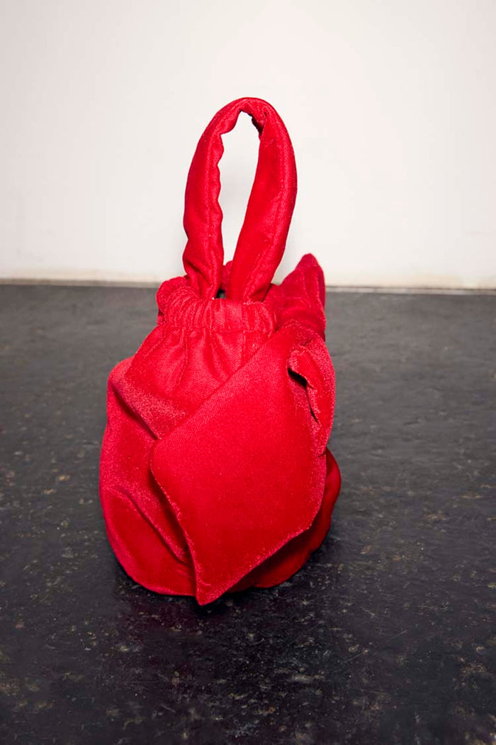 Red Velvet Bow Bag with an oversized bow detail and padded top handle, set against a dark textured floor.