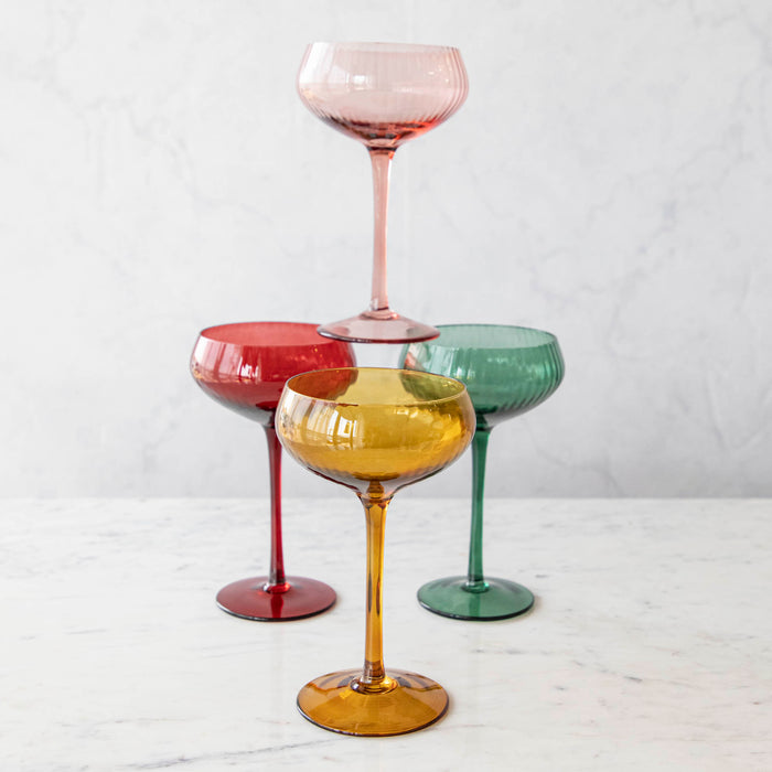 A set of stemmed champagne coupe glasses in various colors including green, pink, red, and gold.