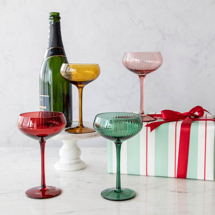 A set of four stemmed champagne coupe glasses in different colors: red, pink, green, and yellow, displayed on a marble surface.