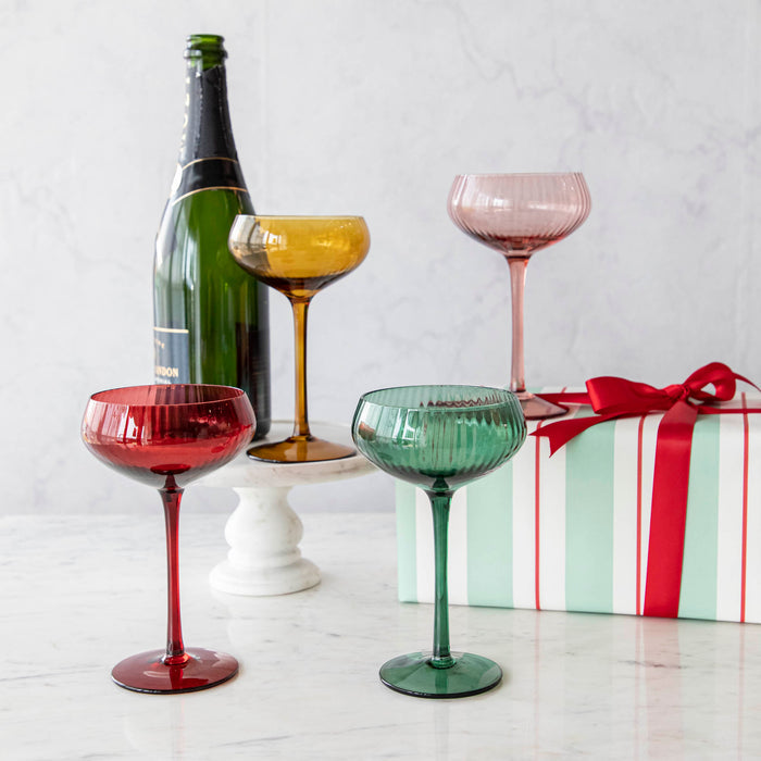 A set of four stemmed champagne coupe glasses in different colors: red, pink, green, and yellow, displayed on a marble surface.