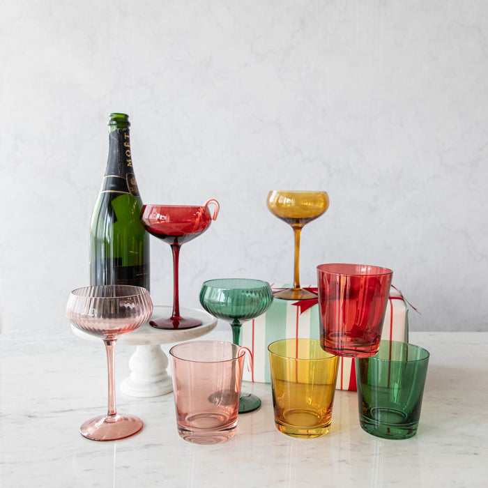 A set of four stemmed champagne coupe glasses in different colors: red, pink, green, and yellow, displayed on a marble surface.