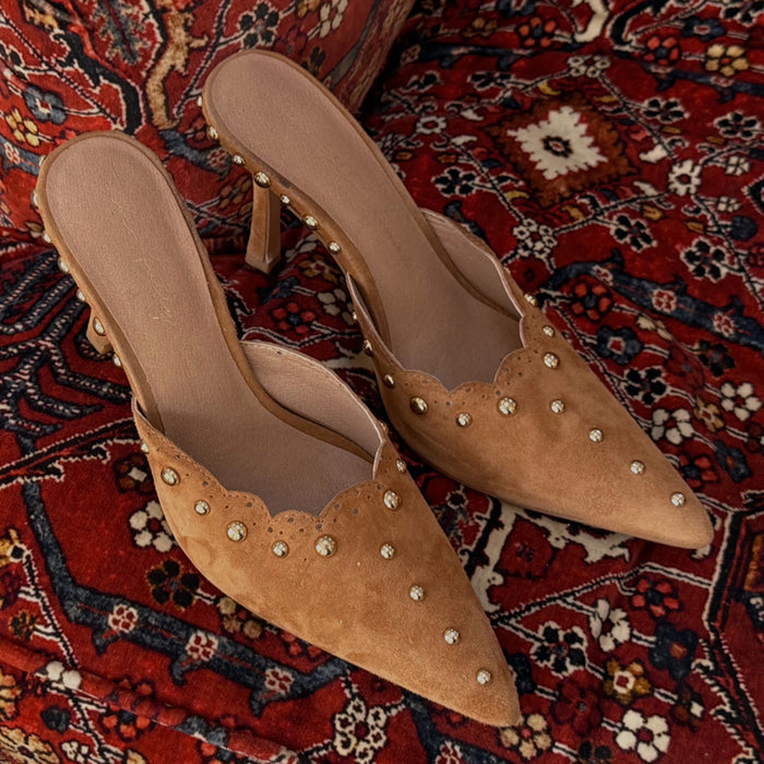 Tan Suede Scallop City Mules with gold studs and a pointed toe, displayed on a red patterned rug.