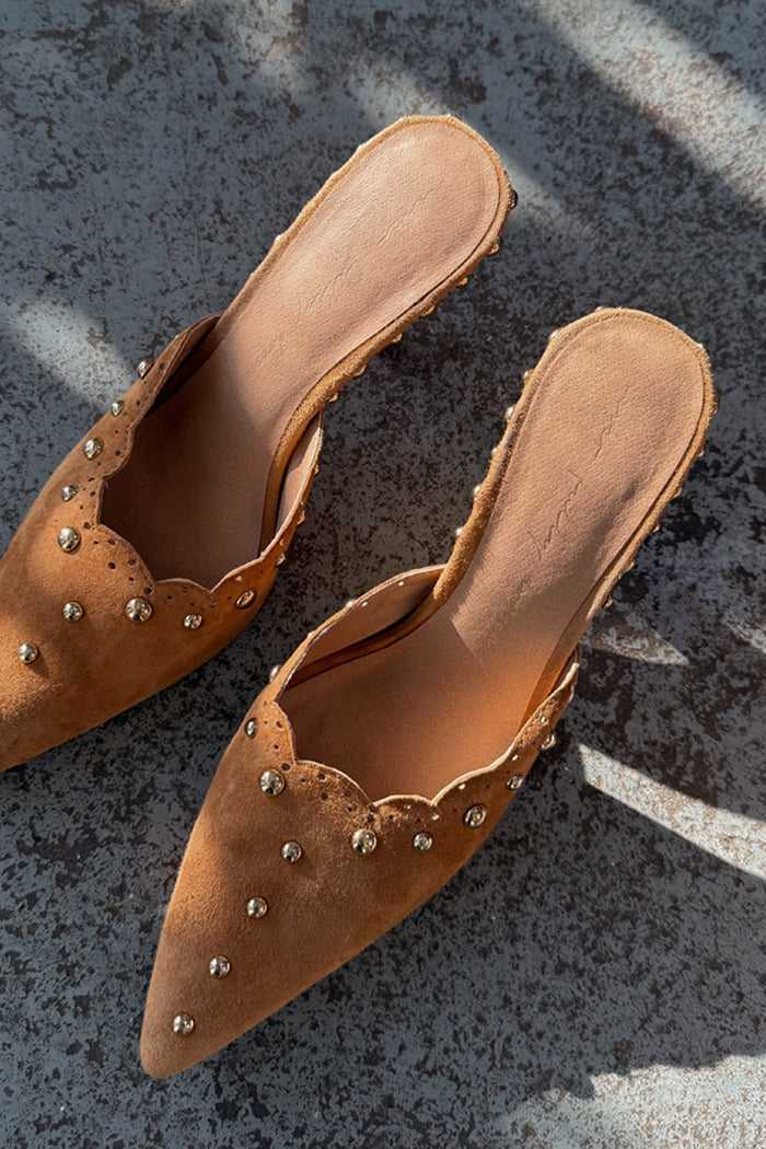Tan Suede Scallop City Mules with gold studs and scalloped edges, shown in a top-down view on a textured surface.