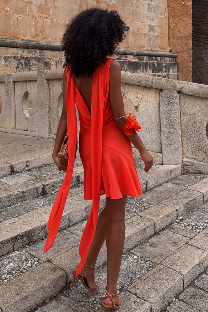 Tomato Mini Sophie Dress in red with a draped back and detachable scarf ties, shown on stone steps.