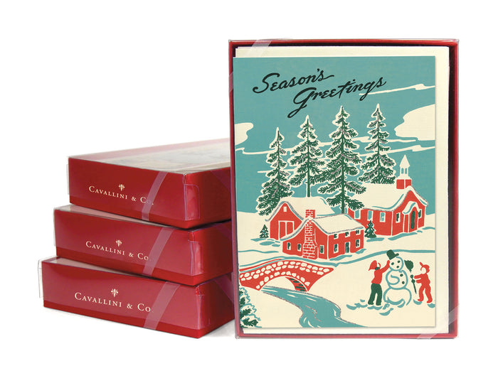 Stack of Season's Greetings Card Sets on their side next to one facing the camera.  Image on card of snowy rural town with children making a snowman
